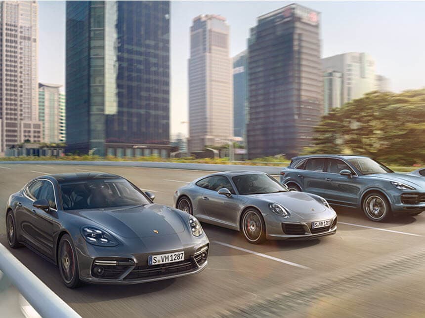 Three silver Porsche cars driving on a city road with tall buildings in the background on a clear day.